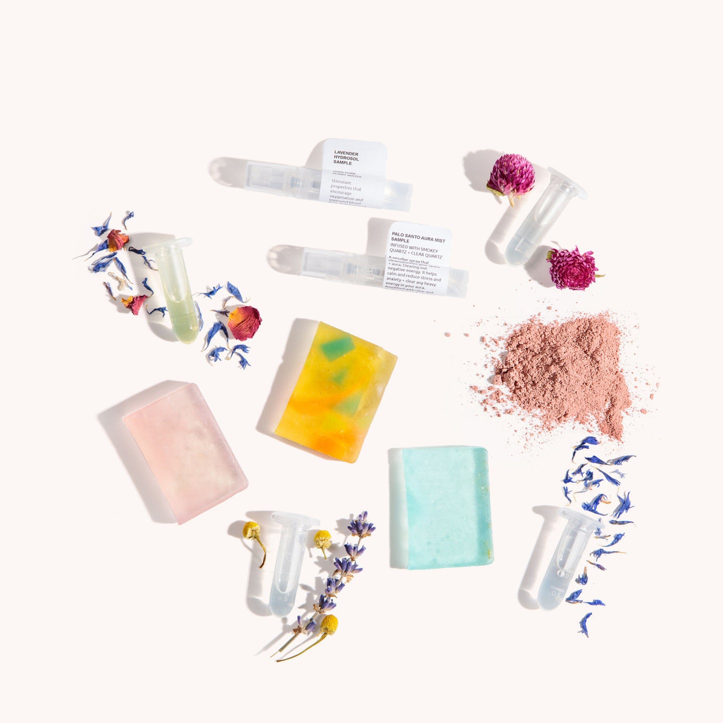 An overhead flat-lay of handmade soap bars in pink, yellow, and mint green, alongside translucent wax melts in tubes, a pile of dusty rose powder, and dried flower petals on a warm off-white background.